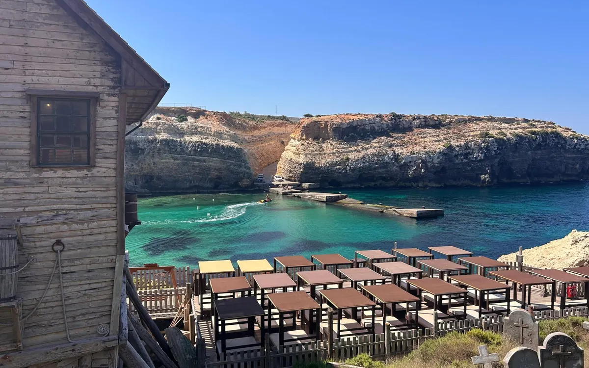 A weathered wooden house above a turquoise bay, rows of shaded day beds on a deck below