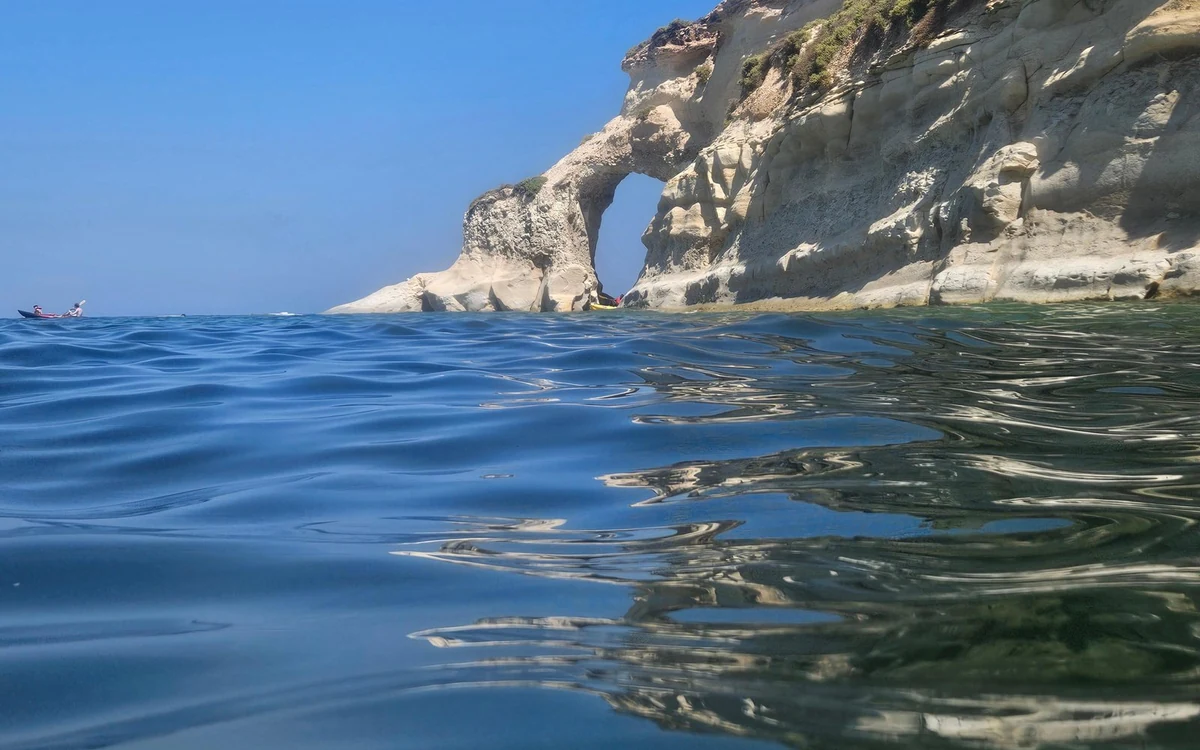 Un arco di roccia alla fine di scogliere bianche visto dal livello dell'acqua, un kayak lontano sulla sinistra