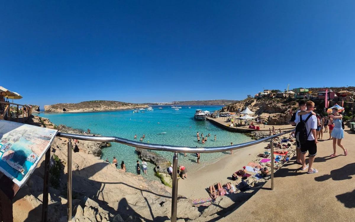 The Blue Lagoon from the cliff path: turquoise water, a sandy beach with sunbeds and a moored tour boat, Malta on the horizon