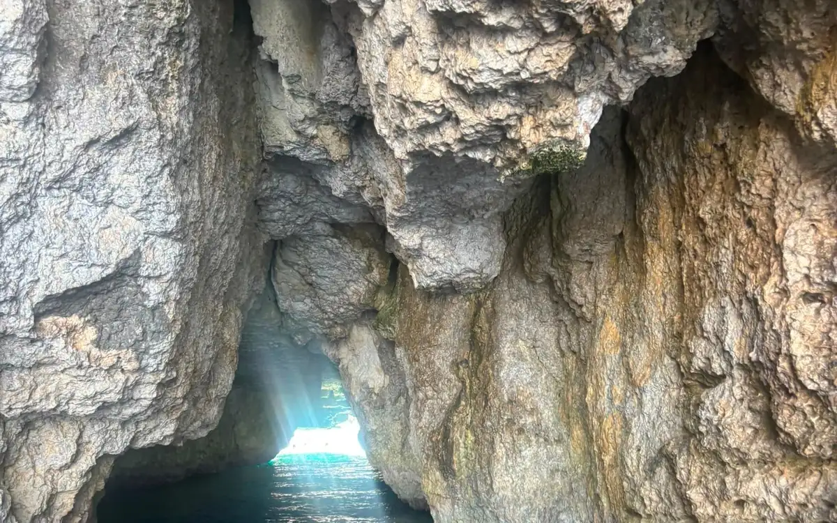 Inside a rock cave seen from a boat, sunlight shining through a narrow opening onto green water
