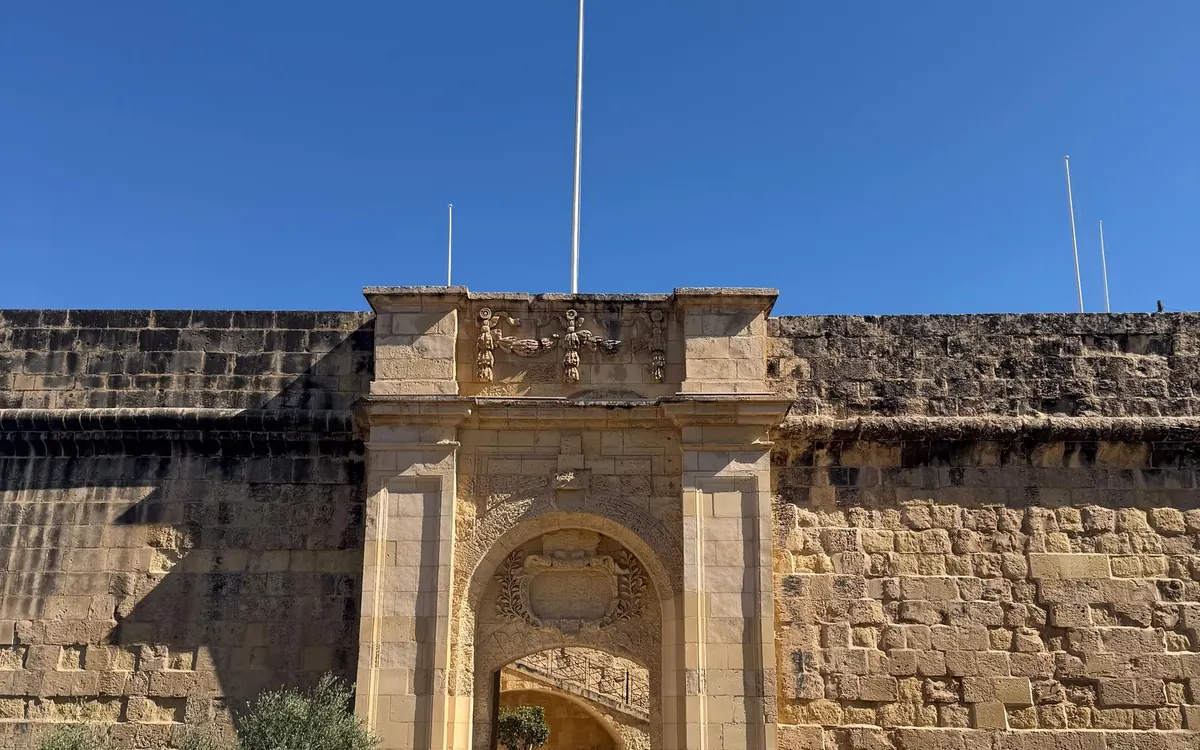 A stone gate with carved trophies above its arch, set in the high walls of a fortified city under a blue sky