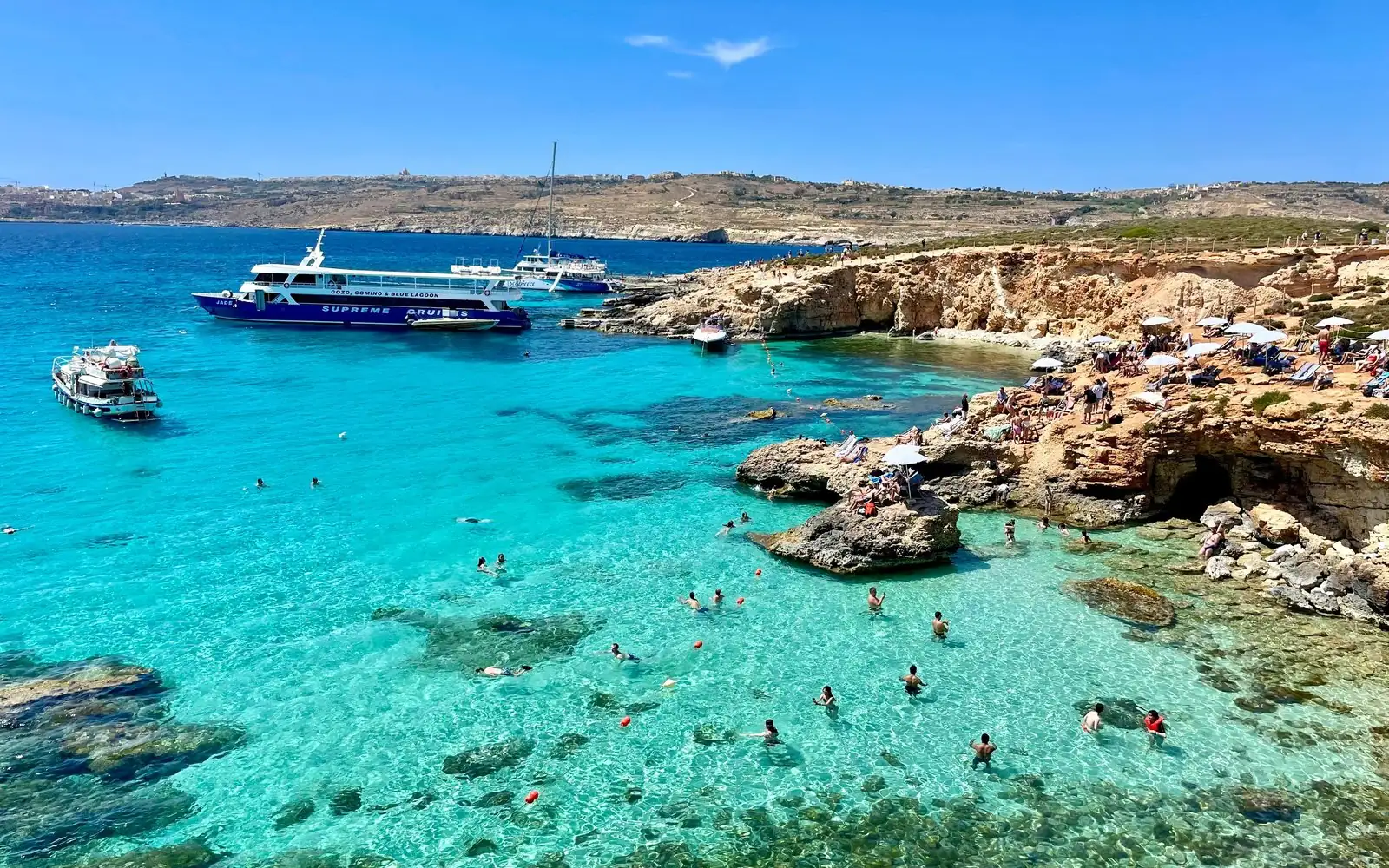 Die Blue Lagoon auf Comino von oben: türkisfarbenes Wasser, Schwimmer, eine felsige Küste und davor vertäute Boote
