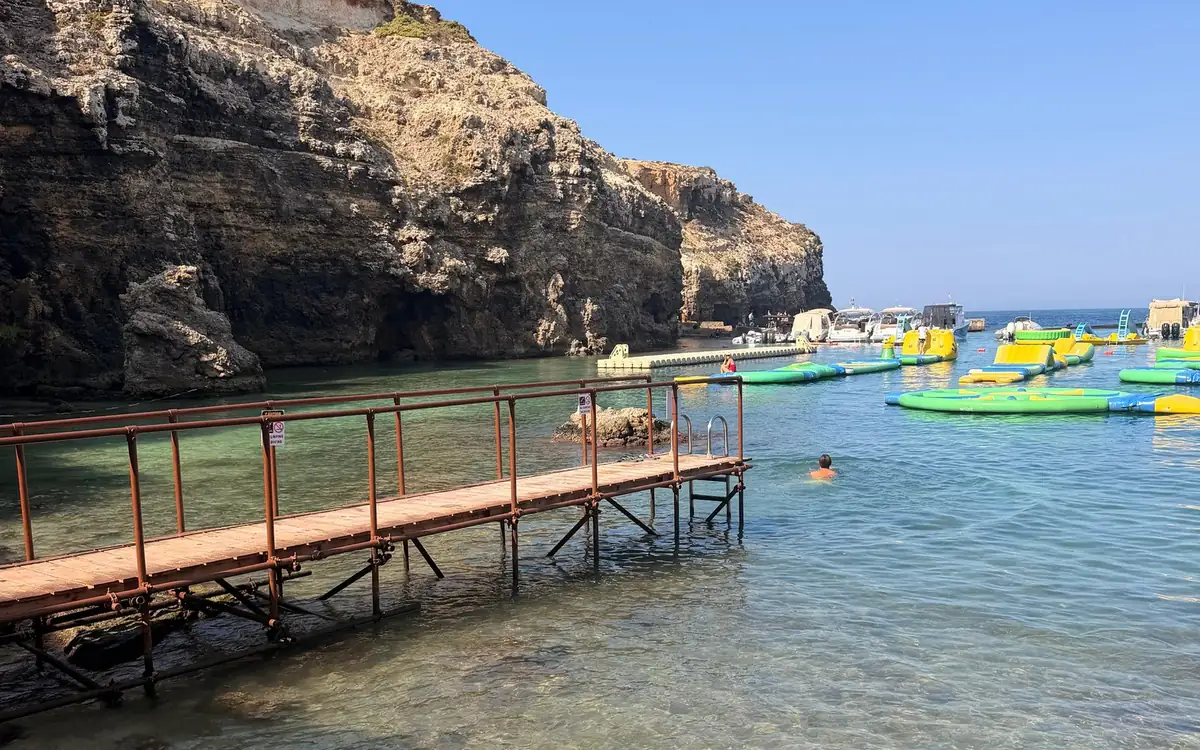 A wooden jetty in clear water below the cliffs, an inflatable water park and moored boats behind