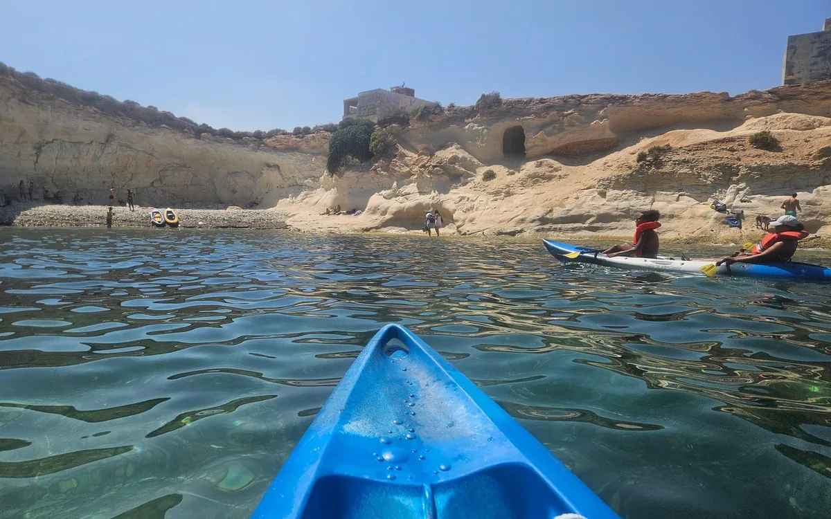 La prua blu di un kayak su acqua verde limpida, due pagaiatori nelle vicinanze e scogliere chiare dietro una spiaggia di ciottoli