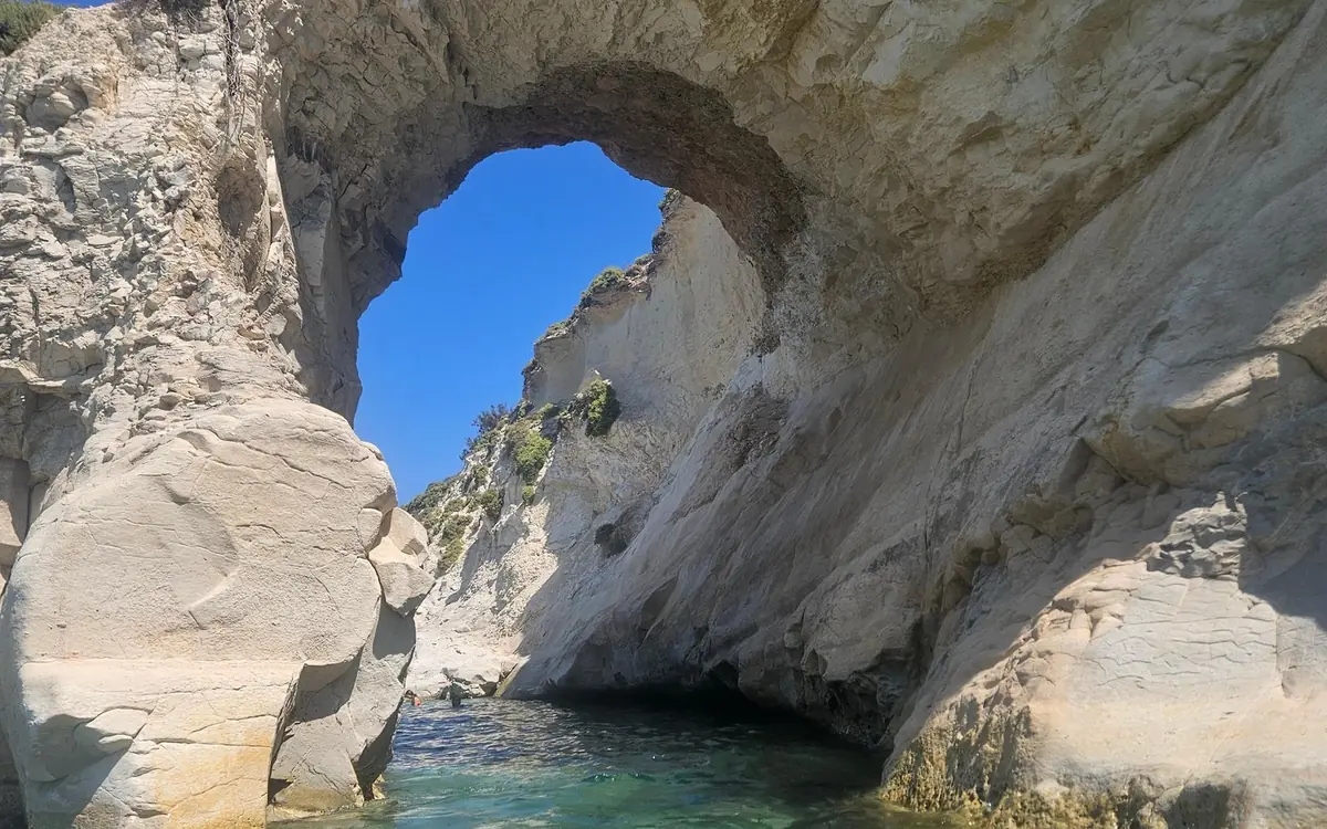 Sguardo verso l'alto attraverso un arco calcareo verso il cielo azzurro, con acqua verde ai suoi piedi