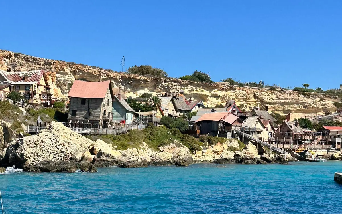 Wooden houses of the Popeye Village film set on the rocks above turquoise water, seen from a boat