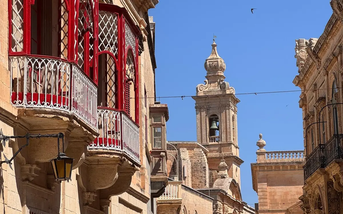 Red wooden balconies on a narrow Mdina street, a church bell tower beyond