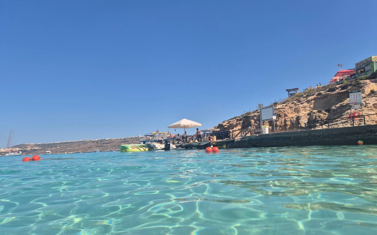 Clear shallow water at the Blue Lagoon, a jetty with moored boats and a beach umbrella, kiosks on the rocky shore above