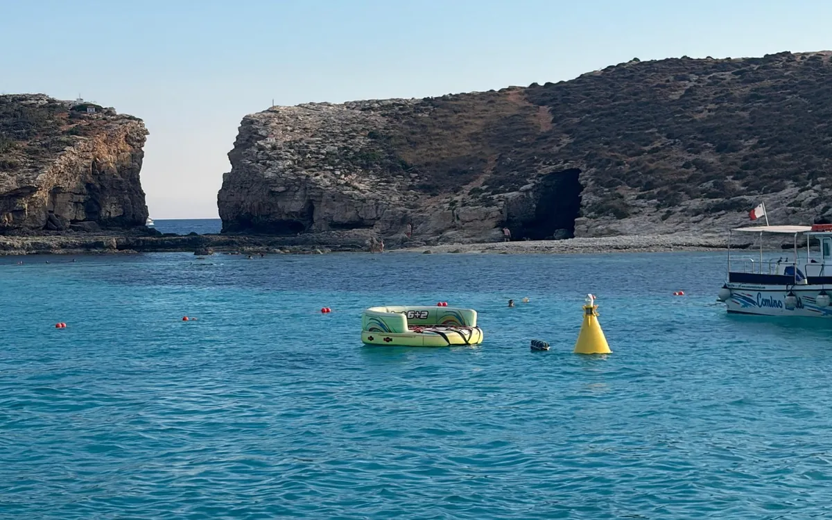 Turquoise water with swim buoys and a moored boat below a rocky islet with a cave