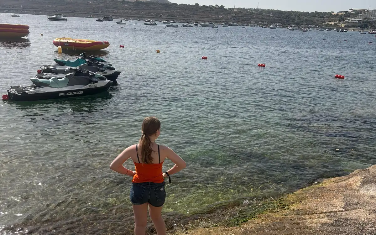 Eine Schwimmerin steht auf den Felsen über klarem Wasser, im Hintergrund vertäute Boote
