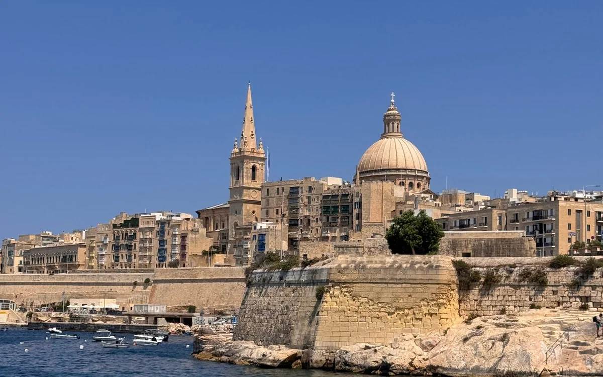 A church dome and a tall spire rising over the bastion walls of Valletta, small boats moored on the water below